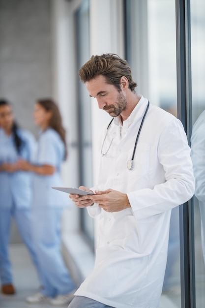 Un médico cansado que usa una tableta digital mientras toma un descanso rápido cerca de una ventana y en un pasillo del hospital durante la pandemia de Covid-19.