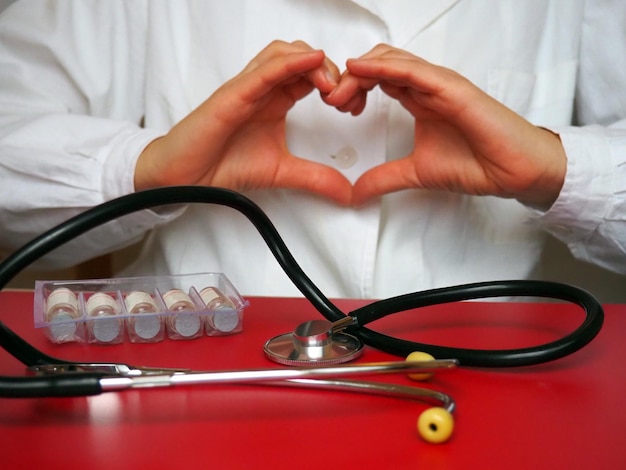 El médico cardiólogo con un estetoscopio muestra el corazón con las manos Estetoscopio y medicamentos sobre la mesa Gesto de las manos Forma del corazón hecha con palmas y dedos Prevención de enfermedades cardíacas