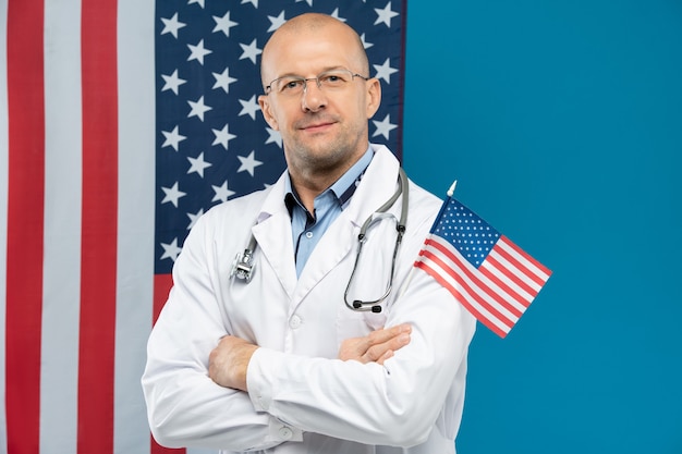 Médico careca de jaleco branco e óculos segurando pequenas estrelas e listras de pé contra a parede com a bandeira americana