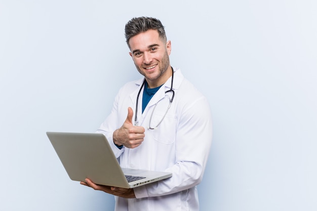 Médico caucasiano homem segurando um laptop sorrindo e levantando o polegar