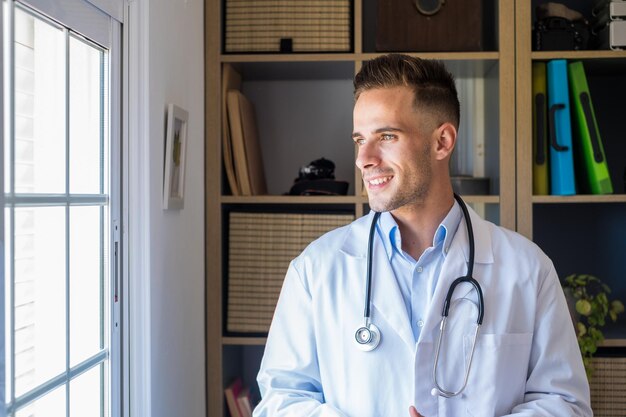 Foto médico caucásico joven y pensativo en uniforme médico blanco mira por la ventana a distancia pensando o reflexionando hombre serio plan de carrera futura o éxito en medicina visualizar en el lugar de trabajo escribiendo xa