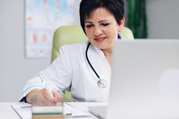 Médico-chefe de meia idade com jaleco branco sentado no local de trabalho, falando com o paciente fazendo videochamada no laptop escrevendo notas no jornal de papel.