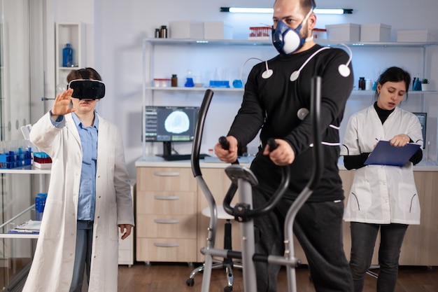 Médico científico en el laboratorio de ciencias del deporte con gafas de realidad virtual mientras el atleta corre, con electrodos conectados al cuerpo que monitorea la resistencia física mientras el escaneo de electrocardiograma se ejecuta en la pantalla de la computadora