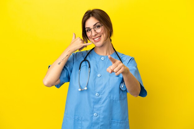 Médico cirujano mujer aislada sobre fondo amarillo haciendo gesto de teléfono y apuntando hacia el frente