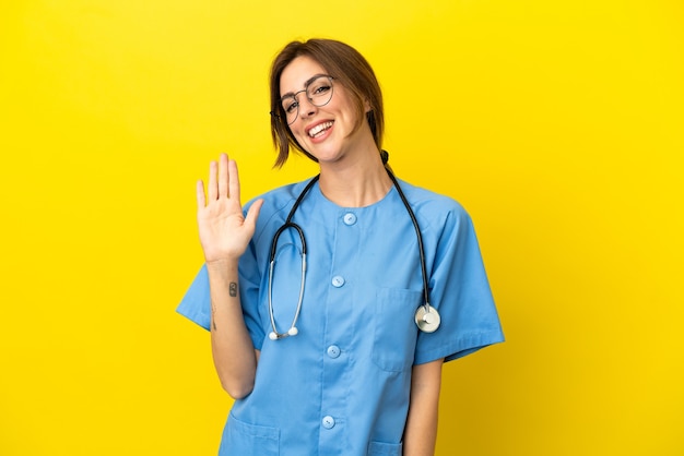 Médico cirujano mujer aislada sobre fondo amarillo saludando con la mano con expresión feliz