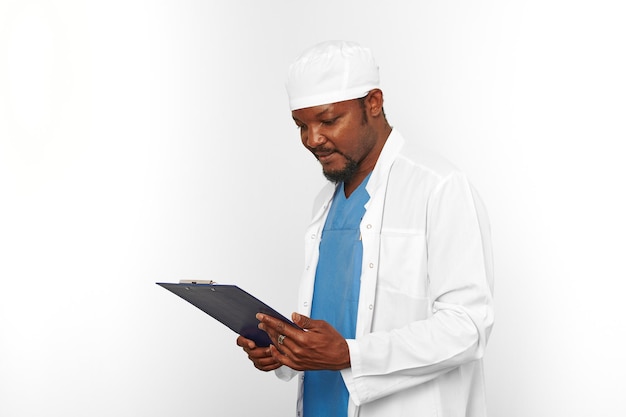Médico cirujano negro hombre barbudo con bata blanca y gorra mira el cuadro médico en el portapapeles en blanco