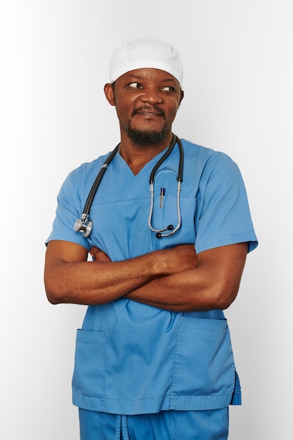 Médico cirurgião negro sorridente homem barbudo de casaco azul e boné médico com estetoscópio de braços cruzados