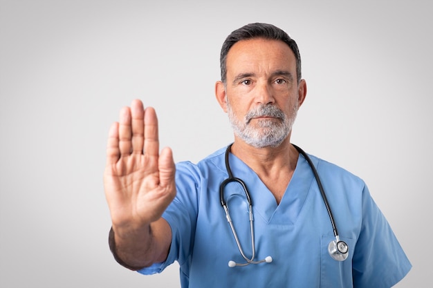 Médico cirurgião sênior caucasiano sério e confiante em uniforme azul fazendo sinal de parada com a mão