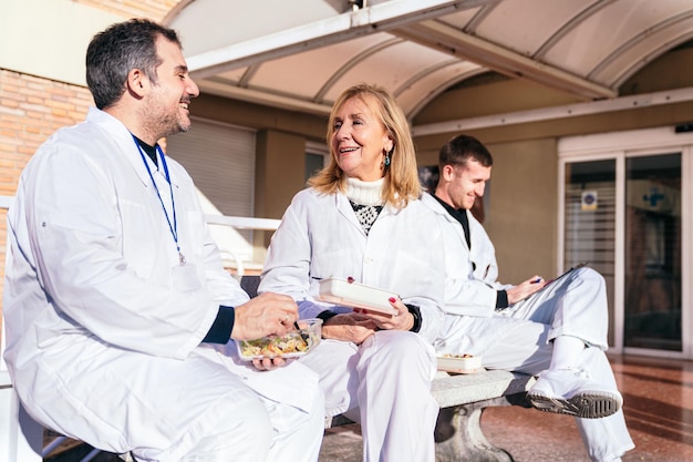 Un médico y un colega alegres sentados al aire libre en un banco disfrutando de una comida saludable