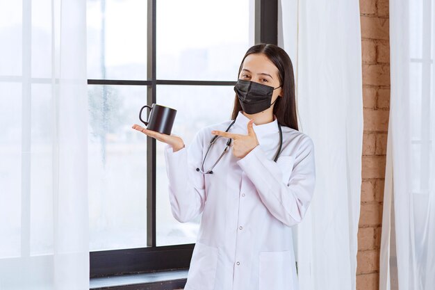 Médico com estetoscópio e máscara preta segurando um copo preto de bebida no intervalo.