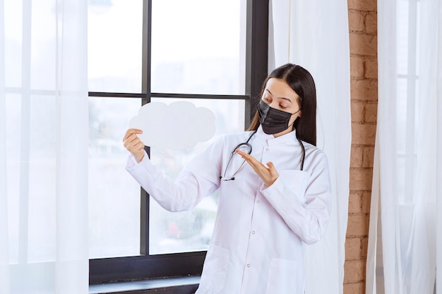 Médico com estetoscópio e máscara preta segurando um quadro de reflexão de forma de nuvem branca.