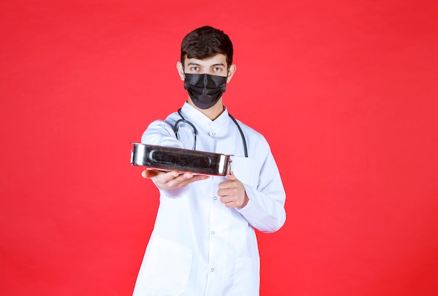 Médico com estetoscópio em máscara preta segurando um recipiente metálico de ferramentas e oferecendo-o ao seu parceiro.