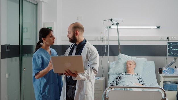 Médico com laptop discutindo com enfermeira antes da consulta com paciente mais velho na cama. Médico consultando mulher sênior com doença para tratamento de recuperação e saúde na enfermaria do hospital