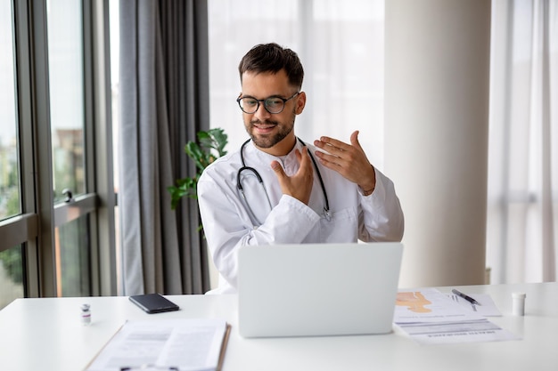 Médico com laptop realiza uma chamada de vídeo on-line e fala com o paciente Médico masculino trabalhando no interior do escritório de mesa em segundo plano