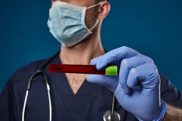 Médico com máscara, luvas descartáveis azuis, traje médico, estetoscópio no pescoço. segurando o tubo de ensaio vermelho de laboratório na mão. posando sobre fundo azul. pesquisa de coronavírus. covid-19. close up, copie o espaço