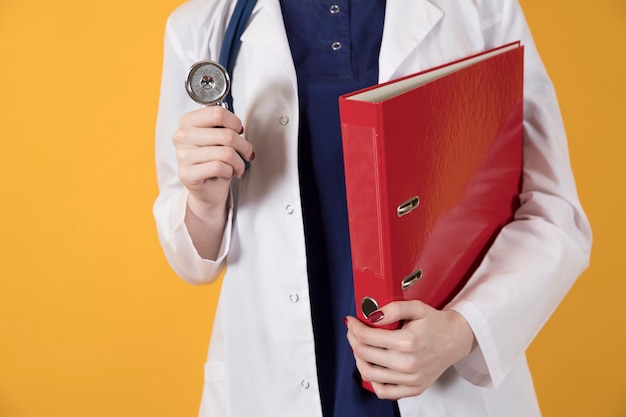 Médico com roupa cirúrgica e jaleco branco segura uma pasta vermelha sobre fundo amarelo