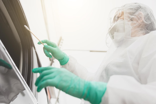 Médico com traje de proteção PPE segurando o kit de teste. Trabalhador médico realizando teste drive-thru Covid-19, coleta de amostra nasal de paciente através da janela do carro, diagnóstico de PCR, kit de teste rápido de antígeno (ATK)