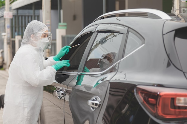 Médico com traje de proteção PPE segurando o kit de teste. Trabalhador médico realizando teste drive-thru Covid-19, coleta de amostra nasal de paciente através da janela do carro, diagnóstico de PCR, kit de teste rápido de antígeno (ATK)