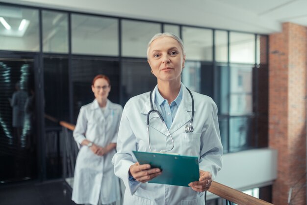 Médico com um estetoscópio. Calma médica sênior com um estetoscópio no pescoço, de pé no corredor ao lado de sua jovem colega