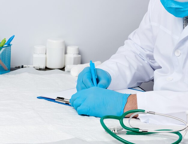 Médico com um jaleco branco médico senta-se à mesa e escreve com uma caneta em uma folha de papel branca