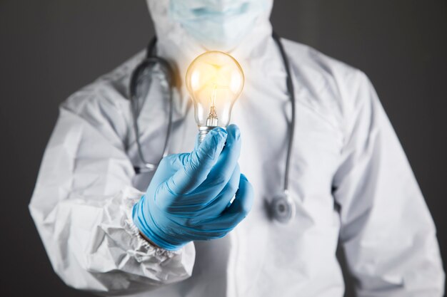 Médico com uniforme de proteção segura uma lâmpada em uma cena cinza