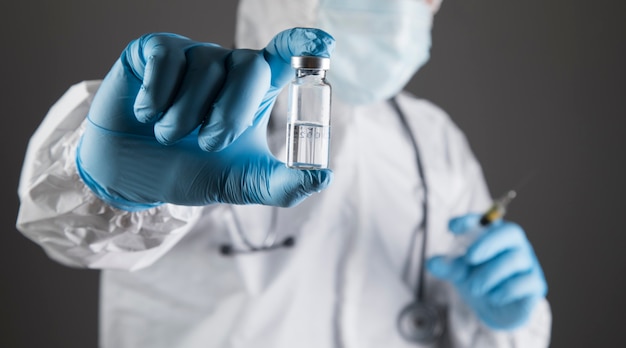 Médico com uniforme de proteção segura uma seringa e uma ampola em uma cena cinza