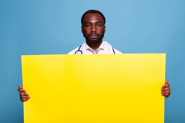 Médico confiante com estetoscópio segurando grande bandeira amarela vazia para conceito médico de cuidados de saúde sobre fundo azul. médico de jaleco branco apresentando painel com espaço de cópia de publicidade.