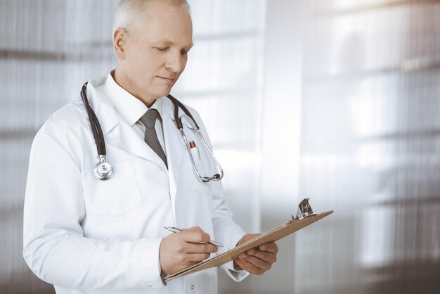 Médico confiante idoso em linha reta e usando a área de transferência na clínica ensolarada. Atendimento médico perfeito. Conceito de medicina.