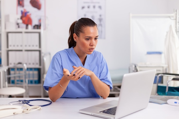 Médico confuso no escritório do hospital enquanto trabalhava no laptop. Profissional de saúde sentado na mesa usando o computador na clínica moderna, olhando para o monitor, medicina.