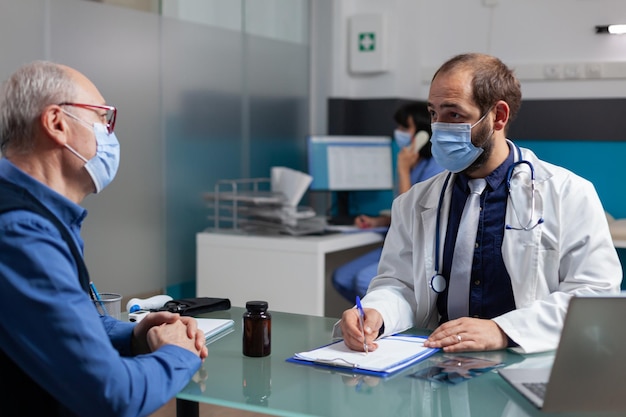 Médico consultando homem maduro e tomando notas na visita de check-up durante a pandemia. Especialista fazendo exame médico com paciente, escrevendo diagnóstico no formulário de consulta no gabinete.