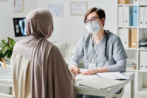 Médico contemporâneo em máscara protetora falando com paciente do sexo feminino em hijab