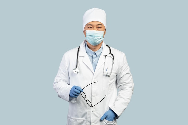 Médico coreano maduro confiante em máscara de luvas protetoras de jaleco branco tira óculos
