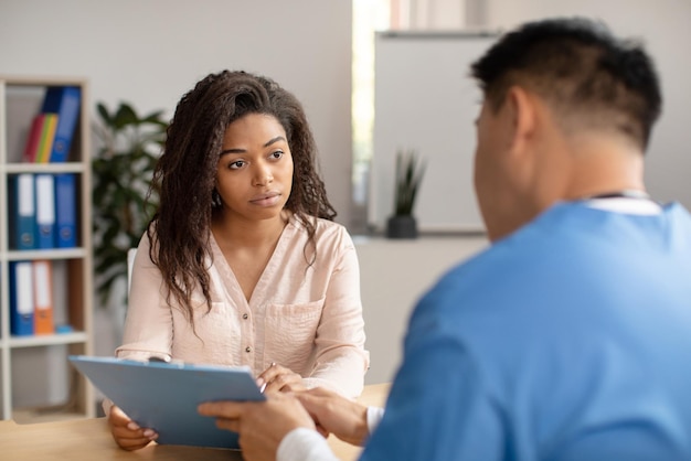 Médico coreano de mediana edad en uniforme consulta a una joven afroamericana triste que muestra una tableta en la clínica