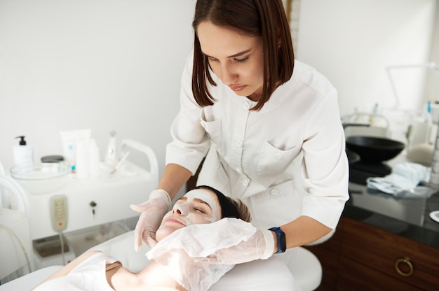 Médico-cosmetologista usando guardanapos para remover a máscara de hidratação de beleza do rosto da mulher em salão de spa. conceito de cuidados com a pele