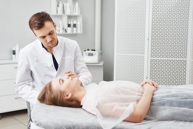 El médico cosmetólogo hace marcas en la cara de una paciente joven que se prepara para el procedimiento de terapia botulínica para las arrugas en un salón de belleza blanco