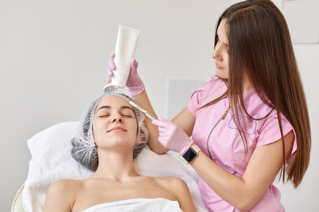 Médico cosmetólogo haciendo masaje de estiramiento facial para joven mujer morena, cliente acostado en los ojos cerrados, con una toalla blanca y gorra médica.