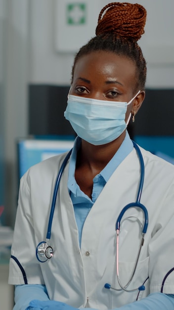 Médico da etnia afro-americana sentado na mesa com laptop e documentos enquanto usava máscara facial e jaleco branco olhando para a câmera. Mulher negra trabalhando como médica no gabinete de saúde