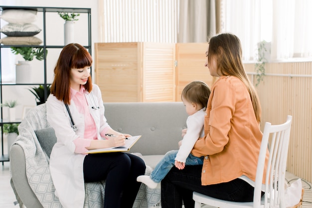 Médico da jovem mulher de jaleco branco escrevendo algo no caderno e mãe com bebê em casa. conceito de medicina, saúde, pediatria e pessoas