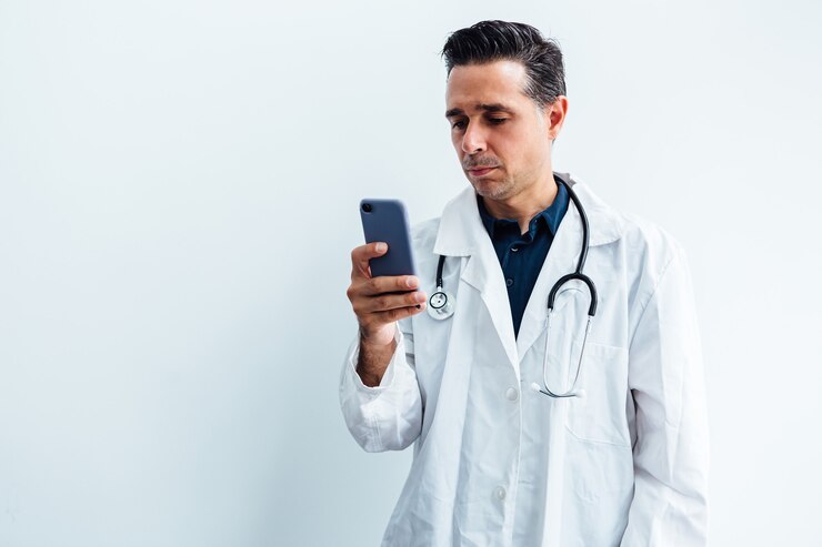 Médico de cabelo preto vestindo jaleco branco e estetoscópio, olhando para seu telefone celular, sobre fundo branco.