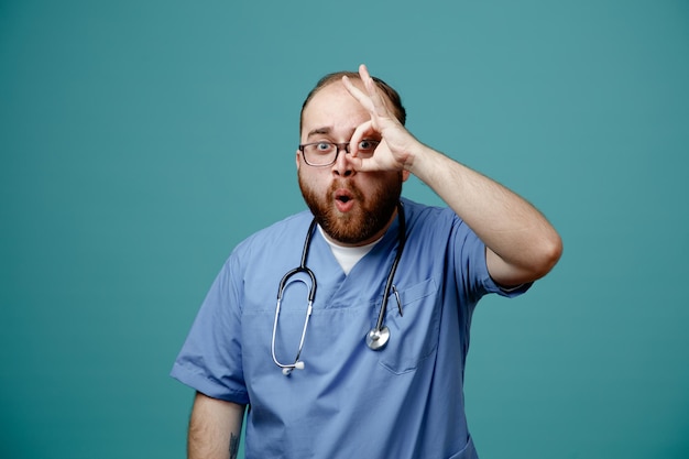 Médico de homem barbudo de uniforme com estetoscópio no pescoço usando óculos olhando por entre os dedos mostrando sinal de ok sendo feliz e surpreso em pé sobre fundo azul