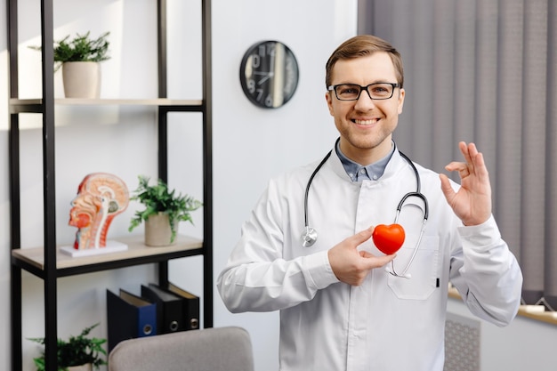 Médico de jaleco branco e óculos segurando um coração vermelho no escritório do hospital Cuidados de saúde médicos e conceito de serviço de equipe médica