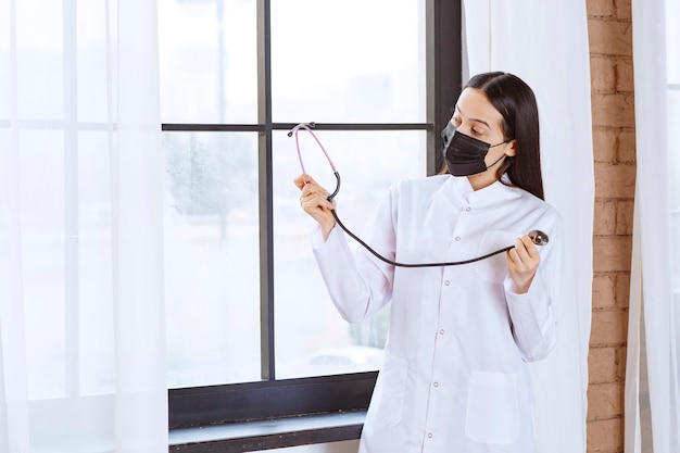 Médico de máscara preta com um estetoscópio em pé perto da janela.