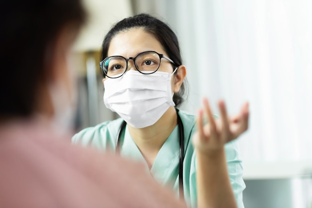 Médico de mulher asiática em óculos de desgaste uniforme verde e máscara cirúrgica falando, consultando e dando conselhos ao paciente do sexo feminino idoso no hospital.