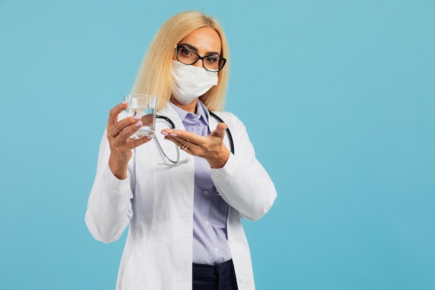 Médico de mulher madura com máscara segura um copo de água e recomenda beber água em fundo azul. conceito covid-19