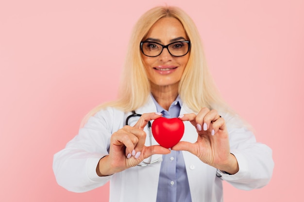 Médico de mulher madura em óculos com estetoscópio segurando coração no fundo rosa com espaço de cópia