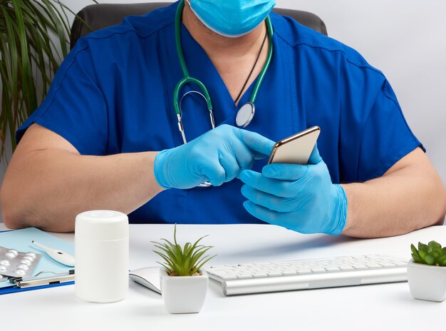 Médico de uniforme azul senta-se em uma mesa e tem na mão um smartphone