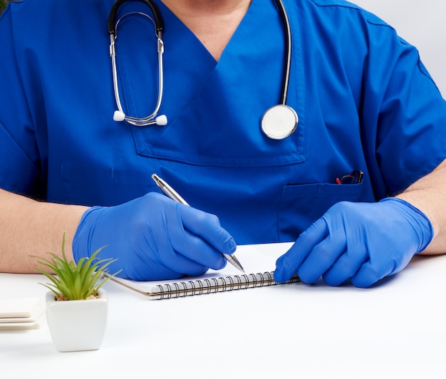 Médico de uniforme azul sentado a uma mesa branca e escrevendo em um caderno de papel