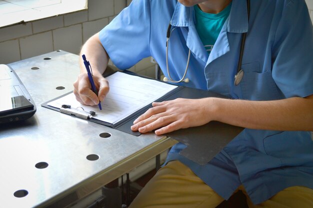 Médico de uniforme azul sentado a uma mesa de metal e escrevendo em um documento