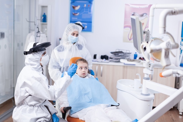 Médico dentista en la consulta de niño pequeño curso en oficina de dentistería. Estomatólogo en traje de protección para coroanvirus como medida de seguridad sosteniendo radiografías de los dientes del niño durante la consulta.