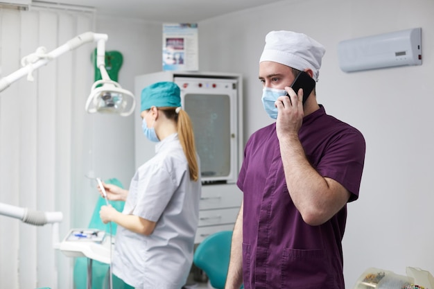 Médico dentista en conversaciones de máscaras médicas en el teléfono móvil de pie en un consultorio dental moderno